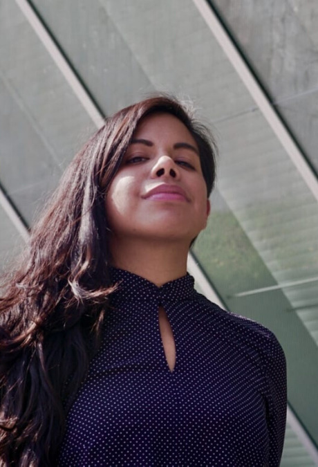 Tania, a Mexican filmmaker with brown skin and long brown hair, peers down at the camera, a small smile on her lips and in her eyes. She wears a dark blue long-sleeved top with small white dots. Behind her, an angular glass building reflects green and whites of the landscape and sun.'